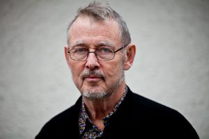 Författaren Lars Ingelstam tittar på betraktaren. Han bär fyrkantiga glasögon, skjorta och kofta och står mot en grå bakgrund.